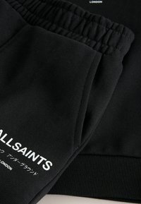 Mustat kangas, jossa on elastinen hihansuu ja valkoinen teksti "ALLSAINTS" sekä japanilaisia merkkejä hihassa tai lahkeessa, näyttäen materiaalin tekstuuria ja ompeluyksityiskohtia.