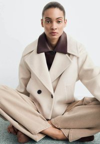 Dwurzędowy, beżowy płaszcz wełniany z ciemno-brązowym kołnierzem, w połączeniu z szerokimi beżowymi spodniami. Model siedzi na jasnoszarej powierzchni.