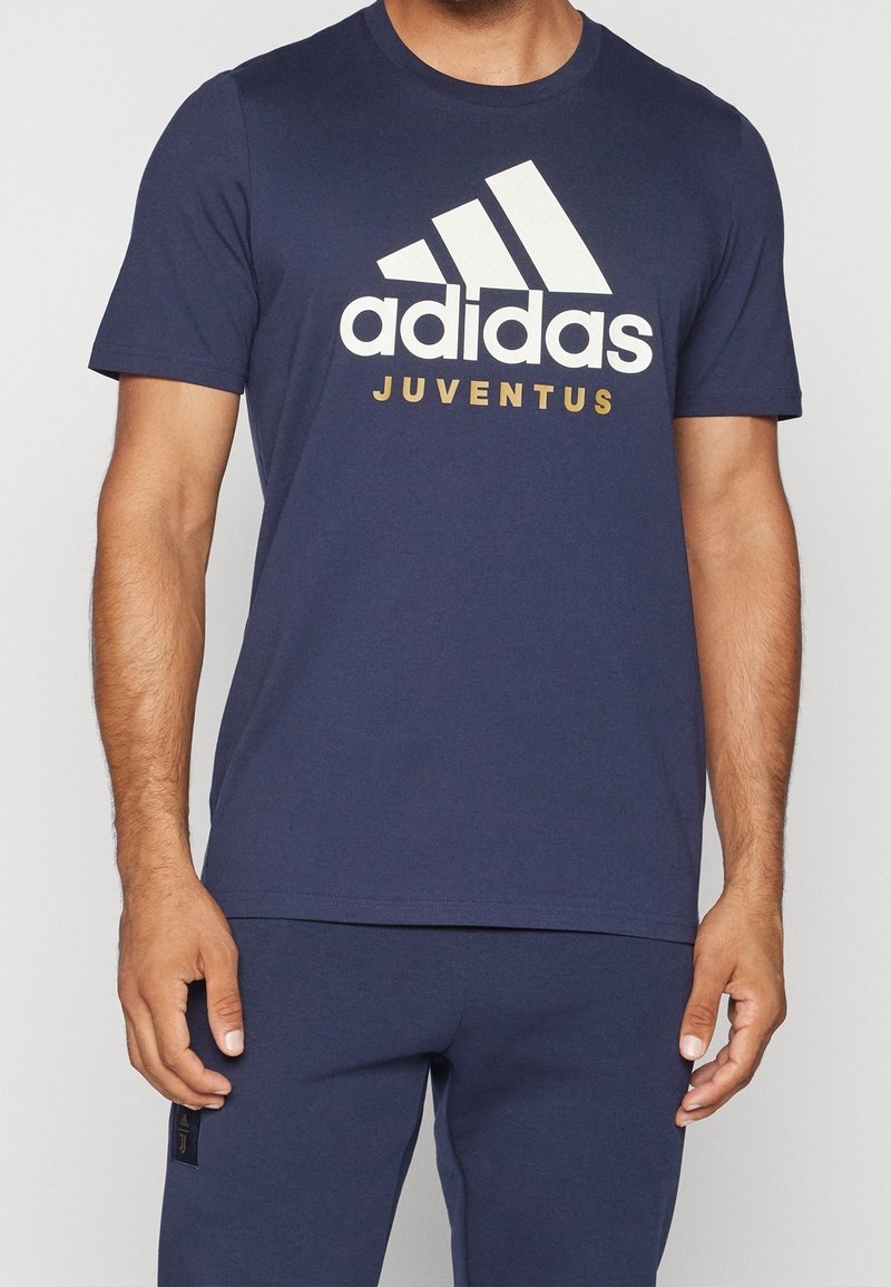 Mężczyzna ubrany w granatowy t-shirt Adidas z dużym białym logo Adidas i złotym napisem "Juventus" na przodzie.