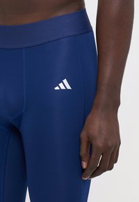 Sötétkék sport leggings sima felülettel, széles derékpánttal és egy fehér Adidas logóval a jobb combon.