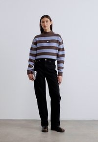 Καφέ και γαλάζιο ριγέ πλεκτό πουλόβερ με ψηλό λαιμό, συνδυασμένο με μαύρο παντελόνι και γυαλιστερά παπούτσια. Το μοντέλο κρατάει ένα μικρό μεταλλικό clutch.