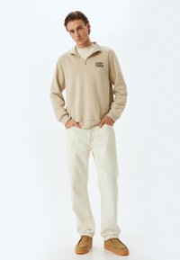 Beige Sweatshirt mit Reißverschluss und dem Text "BEHALTE kreatives DENKEN", kombiniert mit hellen Jeans und hellbraunen Schuhen. Glatte, baumwollähnliche Textur.