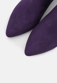 Zapatos de ante púrpura con punta afilada y una textura suave, que presentan un diseño elegante y minimalista, destacando líneas limpias y acentos sutiles.