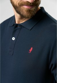 Jūros mėlynos spalvos polo marškiniai, pagaminti iš tekstūruotos medvilnės, su raudonu siuvinėtu logotipu kairiajame krūtinės šone ir trimis baltomis sagomis.