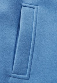 Tessuto blu chiaro con una morbida e leggermente pelosa texture presenta una tasca rettangolare prominente, delineata da cuciture sottili.