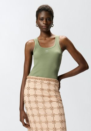 Donna con top verde senza maniche e gonna beige con motivi ricamati a griglia e floreali, in piedi con una mano sul fianco.