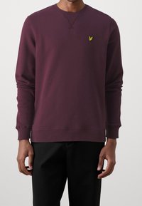 Sudadera burdeos con cuello redondo y puños acanalados. Presenta un pequeño logo amarillo en el pecho. La tela tiene una textura suave. Pantalones negros visibles abajo.