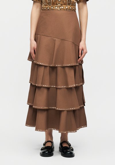 Καφέ midi φούστα με στρώσεις και μικρές λεπτομέρειες από τρουκς στα άκρα κάθε βολάν, συνδυασμένη με μαύρα παπούτσια με λουρί και αγκράφα σε λευκό φόντο.