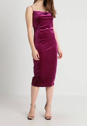 Kvinde iført en ærmeløs, mid-lang lilla fløjls kjole med tynde stropper og sorte åbne hæle med ankelstropper mod en ensfarvet baggrund.