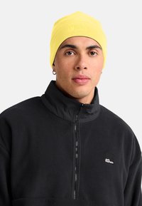 Jonge man die een gele beanie en een zwarte fleece jack met ritssluiting en het Jack Wolfskin-logo draagt, recht vooruit kijkt met een neutrale uitdrukking.