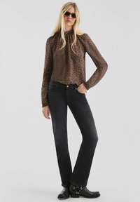 Vrouw in een bruine bloes met lange mouwen, zwarte jeans, zwarte enkellaarsjes en donkere zonnebril, staande met één hand in haar zak.