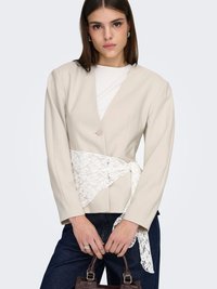 Beige maßgeschneiderter Blazer mit V-Ausschnitt und einem Knopf, akzentuiert durch einen weißen Spitzengürtel. Das Modell trägt dunkle Jeans und hält eine braune Handtasche.