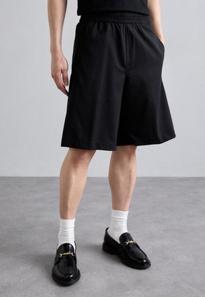 Schwarze Shorts mit elastischem Bund, knielang, aus leichtem Stoff, kombiniert mit schwarzen Slippern und weißen Knöchelsocken.