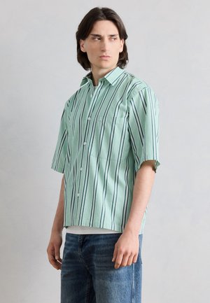 Mann trägt ein kurzärmeliges grünes gestreiftes Hemd mit Knöpfen und blaue Jeans und schaut seitlich vor einfarbigem Hintergrund.