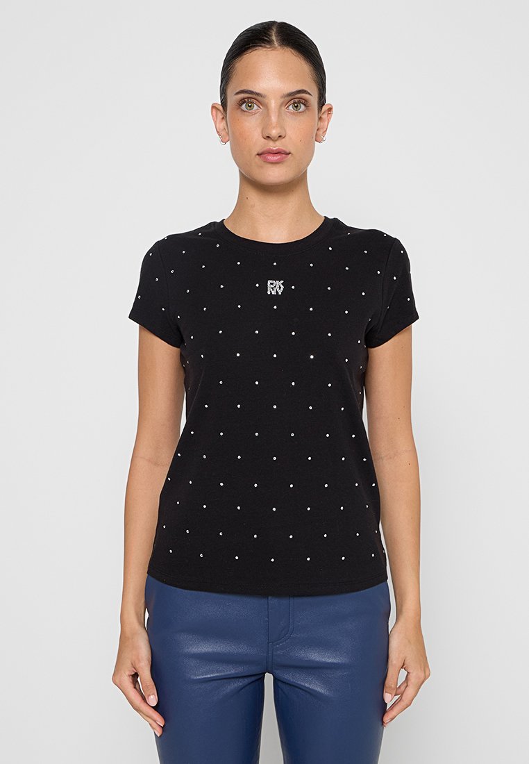 Femme portant un t-shirt noir à manches courtes avec de petits points argentés et un logo "DKNY" argenté, associé à un pantalon en cuir bleu.