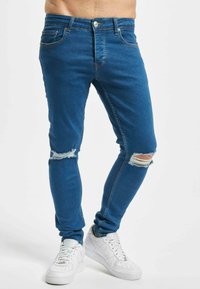 Kék farmer skinny farmernadrág, térdnél szakított részletekkel, öt zsebbel és cipzáros záródással. Fehér sportcipővel viselve.