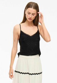 Camiseta de encaje negro con tirantes ajustables, patrón texturizado y escote ondulado, combinada con una falda de cintura alta de color claro.