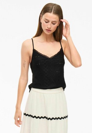 Camisole en dentelle noire avec des bretelles réglables, un motif texturé et un col festonné, assortie à une jupe taille haute de couleur claire.