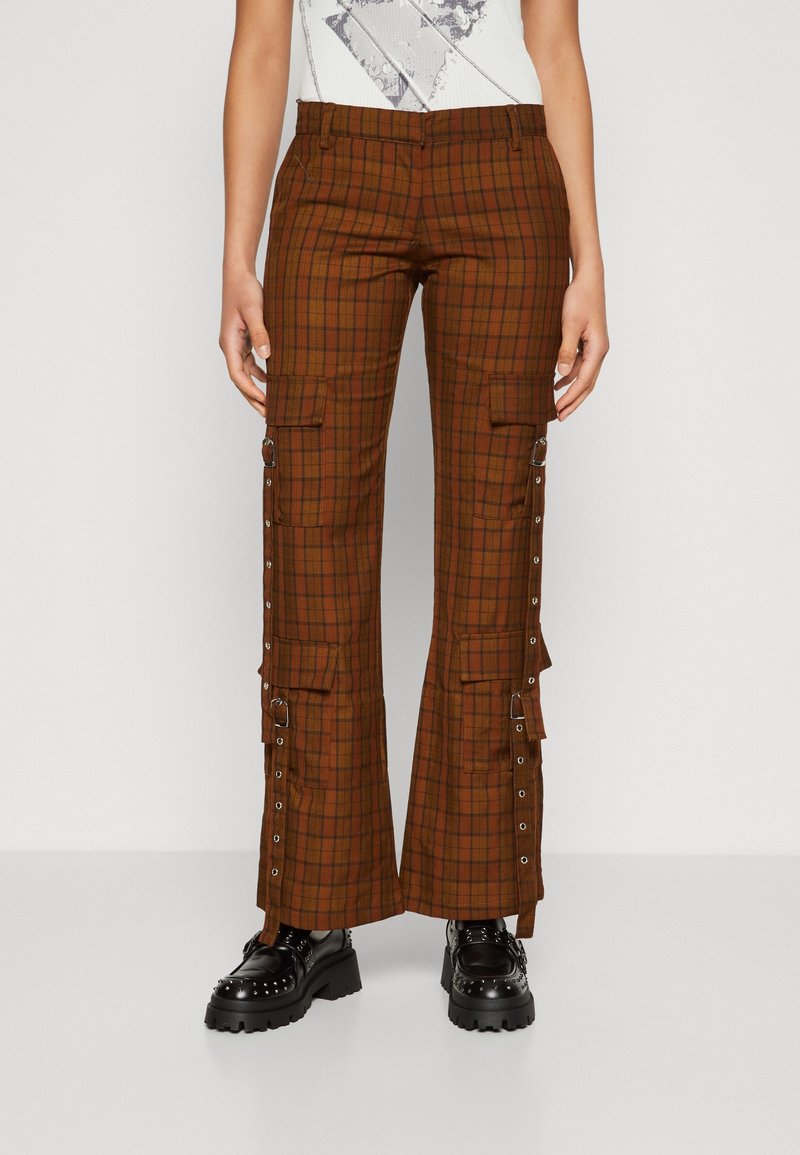 Pantalon cargo à carreaux brun rouille avec poches latérales, œillets en métal et détails à boucle, porté avec des chaussures noires épaisses ornées de clous.