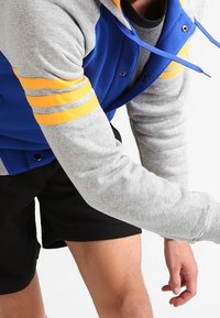 Chaqueta azul y gris con rayas amarillas en las mangas, que presenta botones, cordones y un corte relajado, combinada con pantalones cortos negros.