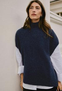 Frau mit langen braunen Haaren, die einen dunkelblauen ärmellosen Pullover über einem weißen Langarmshirt trägt, steht vor einer hellen Wand.