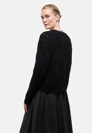 Femme aux cheveux blonds attachés en un chignon bas, portant un pull noir en maille texturée et une jupe plissée sombre, regardant par-dessus son épaule.