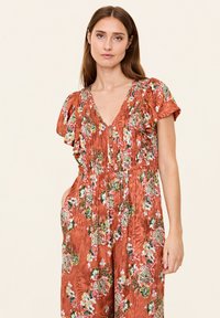 Roodkleurige bloemenoverall met ruches aan de korte mouwen, V-hals, elastische taille en zijzakken. Heeft ingewikkelde bloemenpatronen.