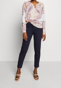 Blouse à manches longues avec un motif paisley en orange, bleu et blanc ; associée à un pantalon slim bleu marine foncé et des sandales à talons carrés beiges.