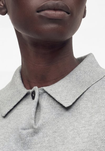Άτομο με σκουρόχρωμο δέρμα που φοράει γκρι ανοιχτό πλεκτό πόλο πουκάμισο με γιακά και ένα μαύρο κουμπί κοντά στον λαιμό.