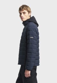 Jeune homme portant une veste matelassée bleu marine avec une capuche, debout de profil devant un fond uni clair.