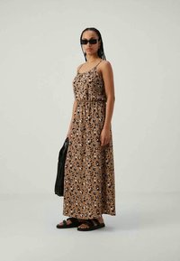 Mulher vestindo um vestido maxi sem mangas com padrão de leopardo castanho, sandálias pretas, óculos de sol escuros e segurando uma bolsa preta em frente a um fundo simples.