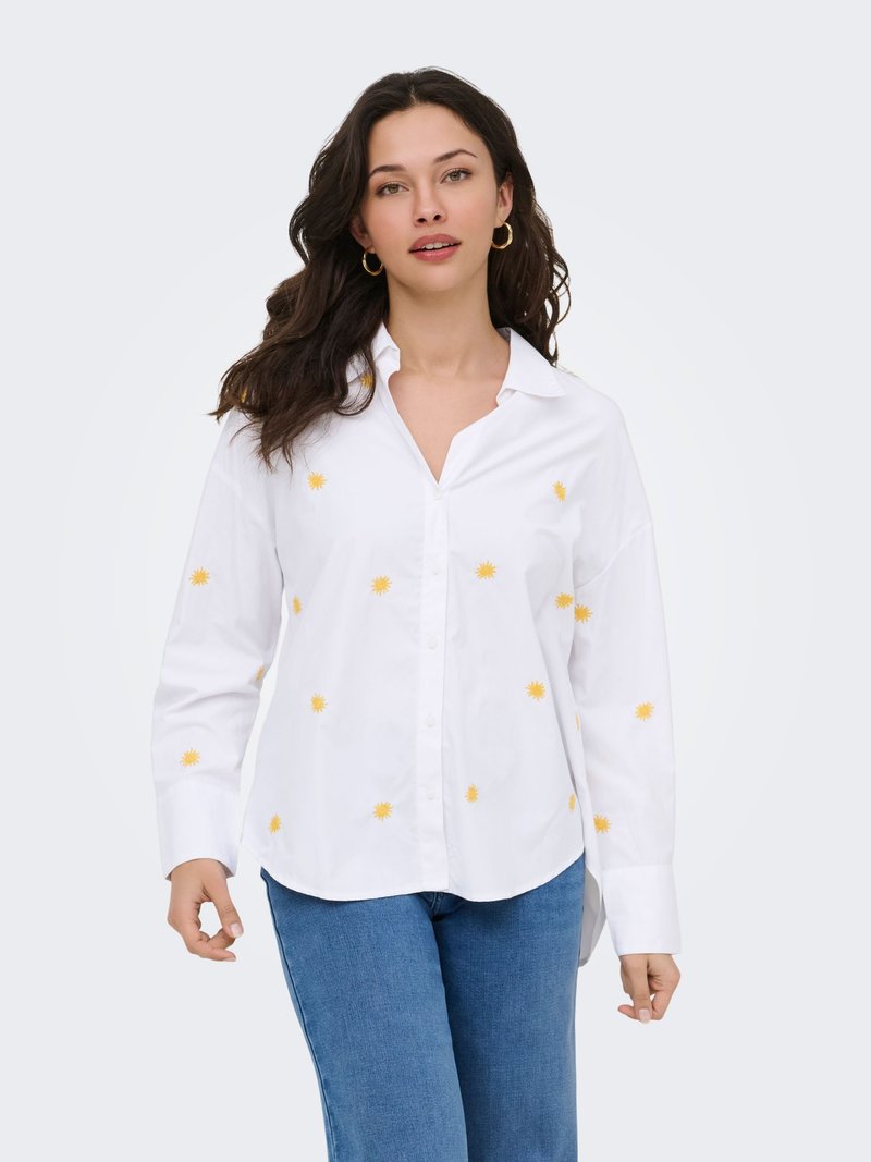 Femme portant une chemise blanche à manches longues à boutons ornée de petits motifs de soleil jaunes et un jean bleu, debout devant un fond uni.