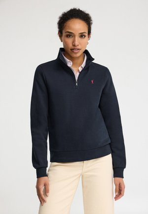 Mujer con un pulóver azul marino de media cremallera sobre una camisa rosa claro con cuello y pantalones beige, de pie contra un fondo blanco liso.