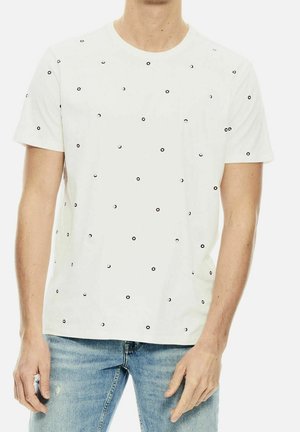 Homme portant un t-shirt blanc à manches courtes avec de petits motifs de cercles noirs et bleus, assorti à un jean bleu clair.