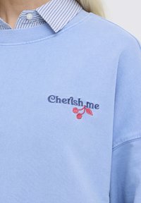 Lichtblauwe sweatshirt over een gestreept overhemd met kraag, met geborduurde tekst "Cherish me" en twee rode kersen op de borst.