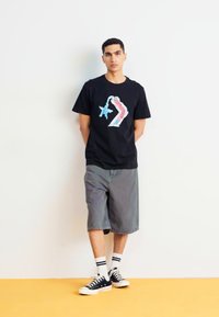 Camiseta negra de algodón con un gráfico colorido en la parte posterior. Combinada con pantalones cortos de mezclilla grises y zapatillas negras con acentos blancos.