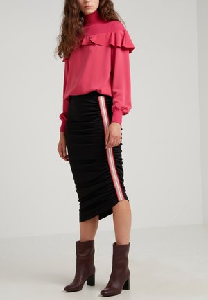Femme portant un pull à col roulé rose vif à volants, une jupe noire froncée avec une bande latérale rouge et blanche, et des bottines marron à talons.