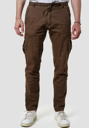 Uomo che indossa pantaloni cargo marroni con vita regolabile tramite coulisse e tasche, abbinati a sneaker bianche, in piedi davanti a uno sfondo neutro.