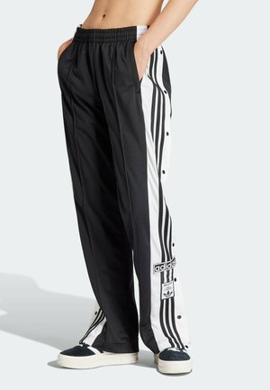 Schwarze Adidas Jogginghose mit weißen Seitenpaneelen, Druckknöpfen, drei schwarzen Streifen, Logos, getragen mit schwarz-weißen Sneakers.