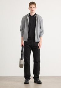 Grijze rits-hoodie over een zwart polo, zwarte jeans en zwarte schoenen. Houdt een grijze schoudertas vast. Minimalistische stijl en neutraal kleurenpalet.