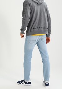 Sudadera gris con capucha y texto negro, combinada con jeans azul claro y zapatillas navy. El fondo es neutro, lo que resalta el atuendo.