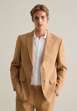 Lichtbruin linnen blazer met notch-lapels, twee knopen en gestructureerde schouders. Gecombineerd met een wit linnen overhemd.