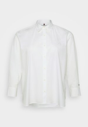 Weißes Langarmhemd mit spitzem Kragen, vorderer Knopfleiste und gesticktem Logo auf der Brust. Glatte Baumwolltextur.