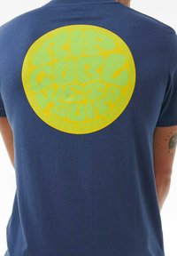 Camiseta azul marino con un gran gráfico circular en la parte posterior. El gráfico presenta texto en amarillo y verde: "RIP CURL WETSUITS." Textura de tela suave.