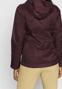 Bordeauxrode waterdichte jas met capuchon, uitgevoerd in een aansluitend model met een gladde stof. Let op de naden en verstelbare manchetten voor een goede pasvorm.