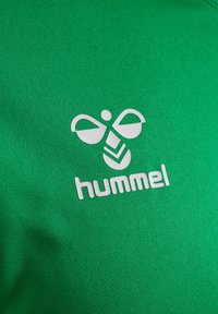 Grøn atletisk skjorte med tekstureret stof, præsenteret med et hvidt Hummel-logo og et særpræget grafisk designelement.