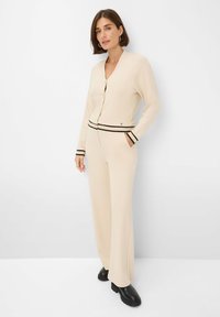 Vrouw die staat met haar handen in de zakken van een crèmekleurige broek, gekleed in een bijpassende cardigan met zwarte strepen op de mouwen en tailleband, zwarte laarzen.