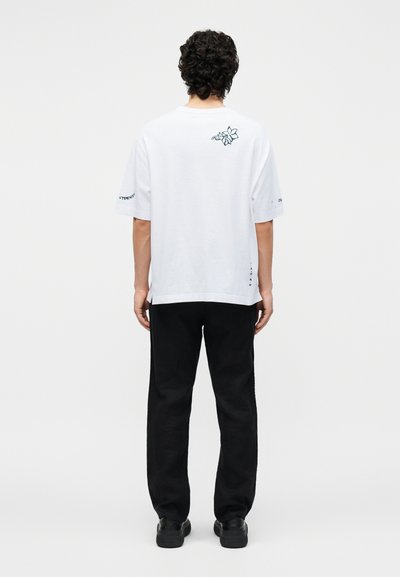 Άνδρας με σγουρά μαλλιά στέκεται με την πλάτη προς το μέρος μας, φορώντας ένα λευκό μπλουζάκι με μικρό λουλουδάτο σχέδιο και μαύρο παντελόνι και παπούτσια.