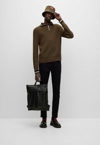 Brown ribbed sweater with a zip collar, black pants, shiny black shoes with red laces, and a black roll-top backpack. Camo bucket hat.