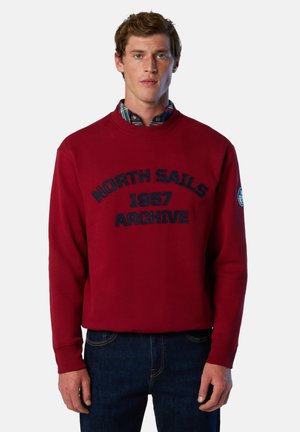 Sudadera roja con cuello redondo, con texto bordado "NORTH SAILS 1957 ARCHIVE" en azul marino. Puños y dobladillo acanalados, llevada sobre una camisa de cuadros.